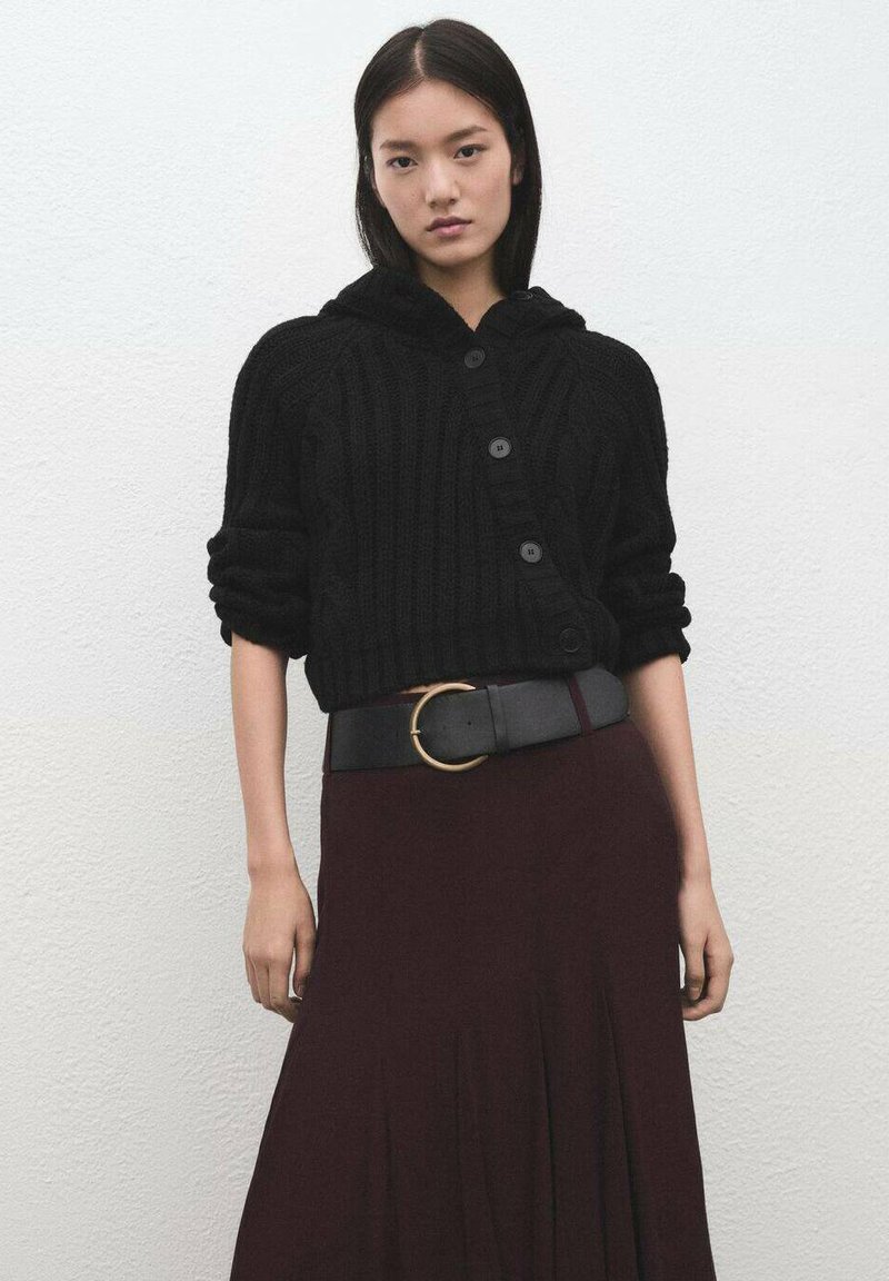 Chaqueta corta de punto negra con capucha, cierre de botones y mangas arremangadas, combinada con una falda burdeos y un cinturón negro con una hebilla de anillo dorado.