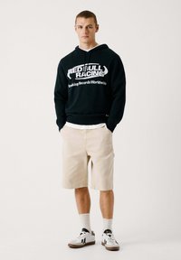 Svart hoodie med vitt "Red Bull Racing"-logotyp, beige shorts, vita strumpor och svartvita sneakers. Enkel och avslappnad design.