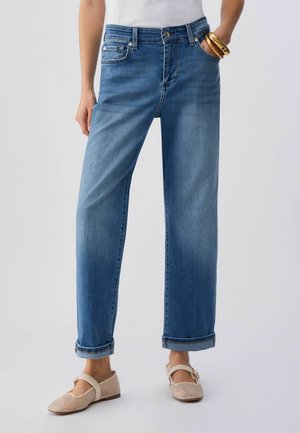 Ženska oblečena v ravne modre kavbojke z zavihki, bež mrežaste Mary Jane čevlje, belo majico in zlate zapestnice na eni roki.