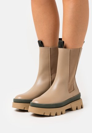 Une paire de bottes en cuir beige mi-mollet avec de grosses semelles crantées tan et des garnitures vertes, portées sur des jambes nues devant un fond blanc.