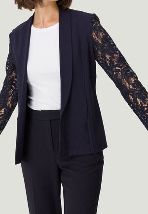 Femme portant un blazer bleu marine foncé avec des manches en dentelle, une chemise blanche côtelée et un pantalon foncé sur un fond clair uni.