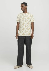 Camiseta de manga corta con fondo beige y patrón de hojas blancas, combinada con pantalones anchos oscuros y zapatos de color claro.