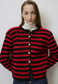 Crvena i crna prugasta kardigan s okruglim izrezom, s zlatnim gumbima duž prednje strane i rebrastom teksturom. Nosi se preko bijele dolčevite.