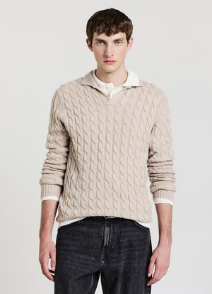 Giovane uomo che indossa un maglione beige a trecce sopra una camicia bianca con colletto, abbinato a jeans neri, in piedi davanti a uno sfondo chiaro e uniforme.