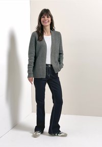 Cardigan gris côtelé porté par-dessus une chemise blanche, associé à un jean foncé et des baskets grises. Debout devant un fond clair.