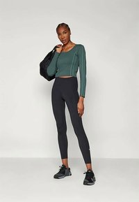 Zöld, hosszú ujjú, cropped felső fehér varrással, fekete, magas derékú leggings-szel és fekete sportcipőkkel párosítva. Nagy fekete táska a vállon.