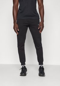 Moški, oblečen v črne tapered trenerke, črne superge Adidas z belimi nogavicami, stoji ob navadni beli podlagi.