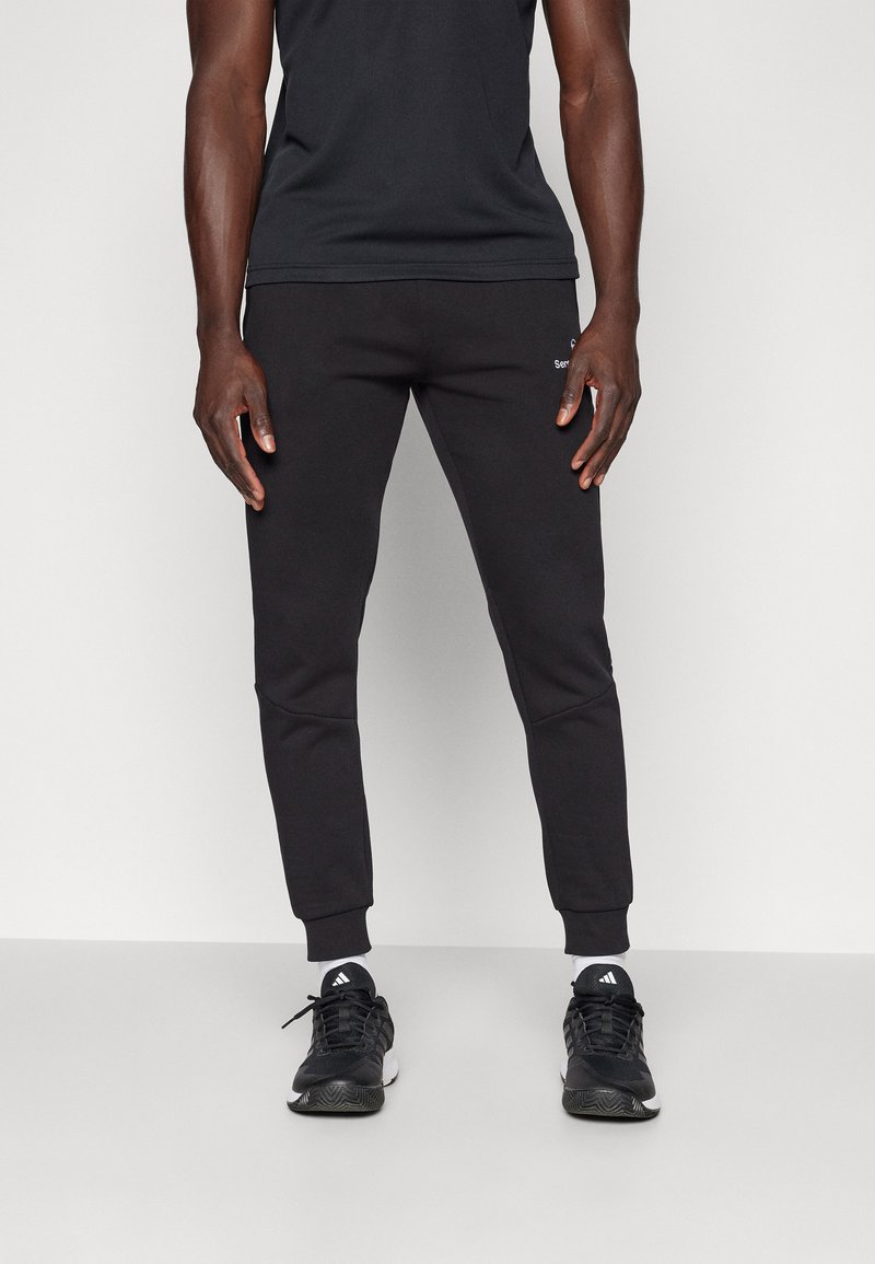 Moški, oblečen v črne tapered trenerke, črne superge Adidas z belimi nogavicami, stoji ob navadni beli podlagi.