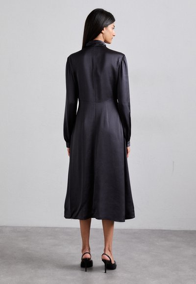 Dunkles Satin-Kleid mit langen Ärmeln, hohem Kragen und figurnaher Passform. Fließt locker bis zur Knielänge. Trägt hohe schwarze Schuhe.