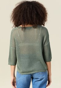 Pull en maille verte avec un tricot lâche et ouvert et des manches trois quarts. Ourlet et poignets côtelés. Porté avec un jean bleu taille haute.