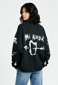 Felpa nera oversize con grafiche bianche, tra cui "Mi Amor", un teschio, frecce e cuori. Materiale in cotone morbido con polsini a coste.