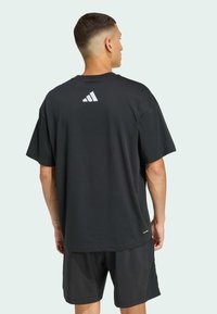 Schwarzes T-Shirt mit kurzen Ärmeln, das auf der Rückseite ein weiß gestreiftes Logo zeigt. Der Stoff wirkt glatt und hat einen lässigen Schnitt. Kombiniert mit dunklen Shorts.