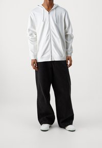 Sudadera blanca con cremallera, capucha ajustable y bolsillos frontales, combinada con jeans negros de corte holgado y zapatillas blancas.