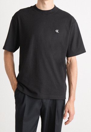 Mann trägt schwarzes Kurzarm-T-Shirt mit kleinem weißem „cK“-Logo auf der linken Brust, kombiniert mit lockerer schwarzer Hose vor einfachem Hintergrund.