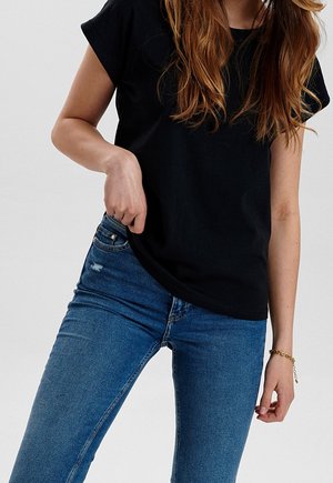 Kobieta o długich rudych włosach, ubrana w czarny t-shirt i niebieskie dżinsy, poprawia koszulkę jedną ręką na tle jednolitego tła.