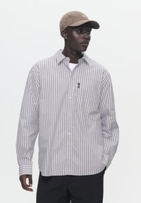 Light gray striped button-up shirt with long sleeves and a pocket. Worn over a white t-shirt, paired with a tan cap.