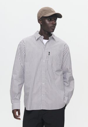 Hellgrau gestreifte Button-Down-Bluse mit langen Ärmeln und einer Tasche. Über einem weißen T-Shirt getragen, kombiniert mit einer beigen Kappe.