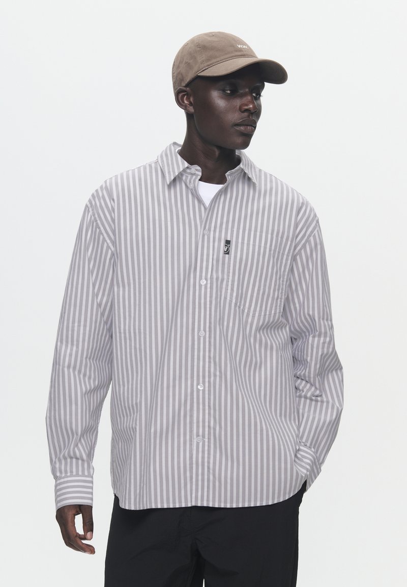 Light gray striped button-up shirt with long sleeves and a pocket. Worn over a white t-shirt, paired with a tan cap.