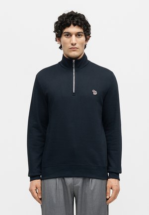 Άντρας με κοντά σγουρά μαλλιά που φοράει ένα μπλε σκούρο πουλόβερ με φερμουάρ μέχρι τη μέση και ένα μικρό πολύχρωμο λογότυπο ζώου, που στέκεται σε απλό λευκό φόντο.