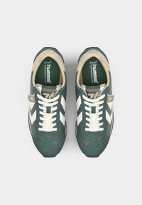 Sneaker verde scuro con strisce a chevron bianche, accenti beige e lacci piatti. Presenta una soletta ortolitica e una suola testurizzata.