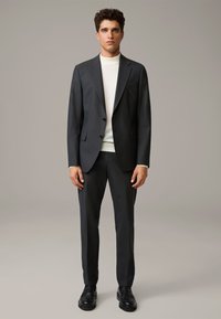 Dark gray tailored suit with a single-breasted jacket, notched lapels, and matching trousers. Worn over a white turtleneck, completed with black shoes.