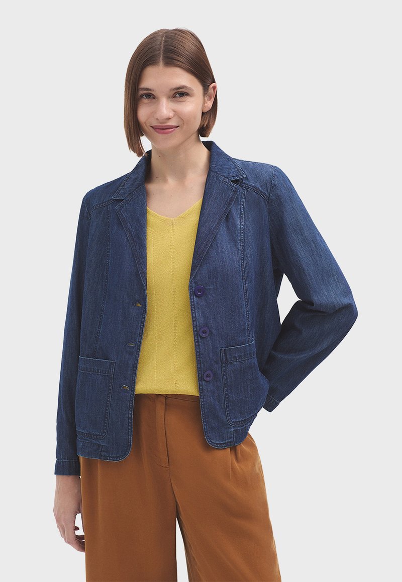 Chaqueta de mezclilla con cuello en pico, dos bolsillos frontales y tres botones morados, llevada sobre una blusa amarilla y pantalones marrones.