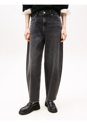 Personne portant un jean noir taille haute coupe ample et des mocassins en cuir noirs, debout les mains détendues le long du corps.