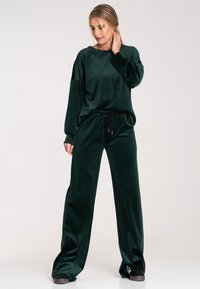 Zielony welurowy dres z długimi rękawami, luźnym krojem, elastyczną talią i kontrastowym sznurkiem. Czarne trampki dopełniają całość.