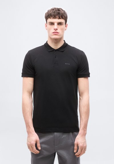 Jeune homme portant un polo noir BOSS et un pantalon gris, debout devant un fond uni gris clair.