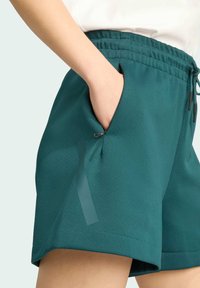 Teal athletic shorts with an elastic waistband, side pockets, and a smooth texture. Features a subtle design element on the side.