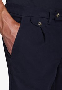 Chinos blu navy con tessuto testurizzato, tasche anteriori e dettagli dei bottoni visibili. Vita cucita, vestibilità sartoriale e finitura liscia.