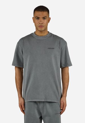 Młody mężczyzna zwrócony przodem, ubrany w luźny szary t-shirt i dopasowane spodnie z małymi czarnymi napisami na klatce piersiowej i udzie.