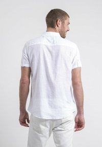 Chemise blanche à manches courtes avec une coupe décontractée, présentant une texture subtile et un col, vue de dos sur un fond neutre.