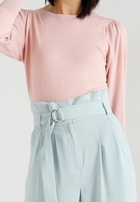 Pull rose clair à manches longues avec épaules bouffantes, associé à un pantalon taille haute bleu clair doté d'une large ceinture et d'une boucle argentée en forme d'anneau.