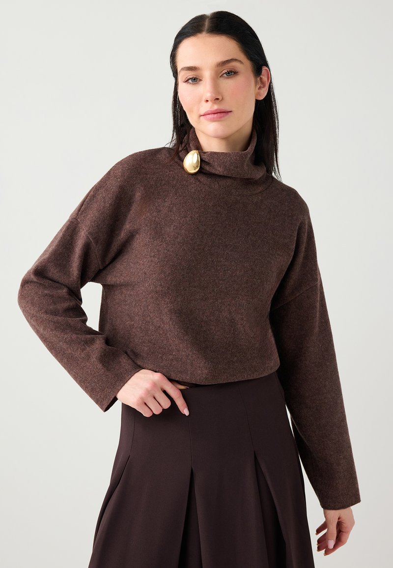 Pull court marron avec un col montant, fabriqué en maille douce. Il présente une broche dorée en accent et des manches amples.
