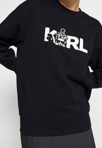 Felpa nera con collo a giro, caratterizzata da una grande grafica bianca che include un testo stilizzato "KARL" e un design di un gatto in stampa leopardata.