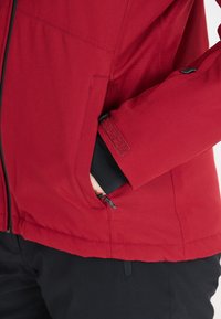 Röd sportjacka med en texturerad yta, med svarta detaljer, dragkedjefickor och justerbara manschetter för en säker passform.