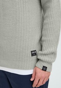 Lichtgrijze gebreide trui met geribbelde textuur, voorzien van zwarte logopatches. Mouwboorden en gelaagd met een witte onderzoom.