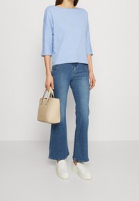 Top in cotone celeste, maniche a 3/4, abbinato a jeans blu a zampa d'elefante. Il modello porta una borsa a tracolla in pelle beige e delle scarpe bianche senza lacci.