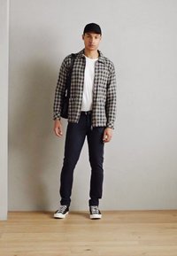 Jaqueta xadrez preta e cinza, camiseta branca, jeans escuros e ténis preto e branco. Modelo está em pé com um boné preto e uma mala.