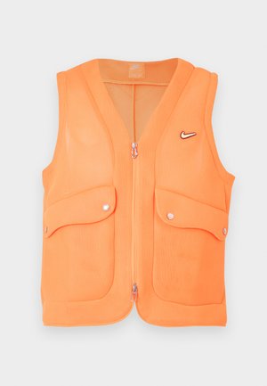 Jeune homme portant une veste zippée orange avec deux poches à rabat, sur un t-shirt noir et un pantalon vert olive, debout devant un fond uni.