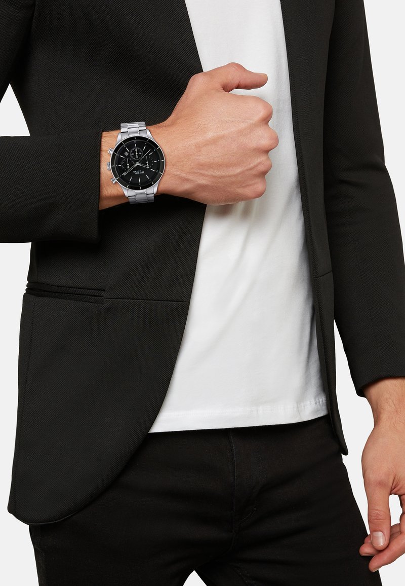 Homme en blazer noir et chemise blanche montrant une montre-bracelet en argent avec un cadran noir au poignet gauche contre un fond blanc.