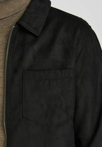 Schwarze Wildlederjacke mit Kragen, Reißverschluss vorne und einer einzelnen Brusttasche. Strukturierter Stoff mit matter Oberfläche, getragen über einem beigen Pullover.