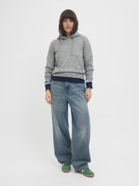 Hoodie gris avec cordon de serrage, poignets côtelés et ourlet bleu marine, associé à un jean bleu ample et des baskets vertes. Fond uni.
