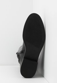 Černá kožená bota s texturovanou gumovou podrážkou; vybavena bočním zipem a blokovým podpatkem pro stabilitu. Zobrazeno z úhlu podrážky.