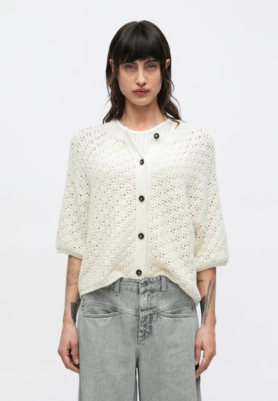 Cardigan en maille couleur crème à manches courtes, avec des motifs ajourés et des boutons noirs, associé à un jean gris taille haute.
