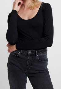 Mustapaita pitkillä hihoilla ja avonaisella kaula-aukolla, yhdistetty tummanharmaisiin korkeavyötäröisiin farkkuihin, joissa on nappi ja etutaskut.