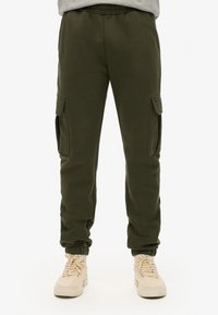 Pantalones cargo verde olivo hechos de un material suave, con bolsillos laterales, cinturilla elástica y puños. Combinados con zapatillas beige claras.