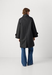 Black waterproof coat with a high collar, wide sleeves, large brown buttons, and a relaxed fit, paired with blue flared jeans.