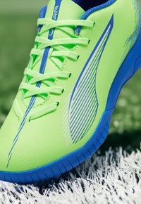 Zapatilla deportiva verde lima con acentos azules, textura de malla y diseño de rayas en el lateral. Presenta cordones planos y una suela de goma.